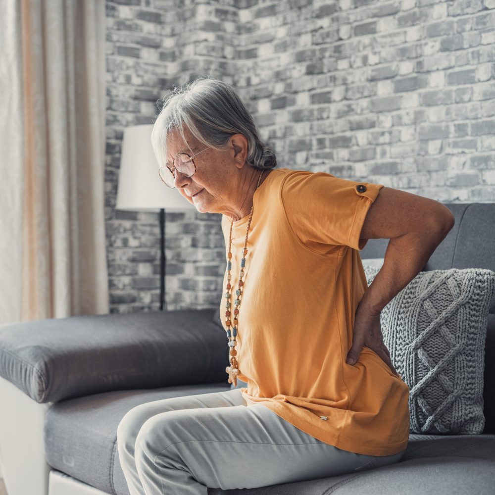Unhappy mature woman touching back, feeling pain, sitting on couch in living room, unhealthy middle aged senior female suffering from backache, spinal problem, rubbing stiff muscles or kidney